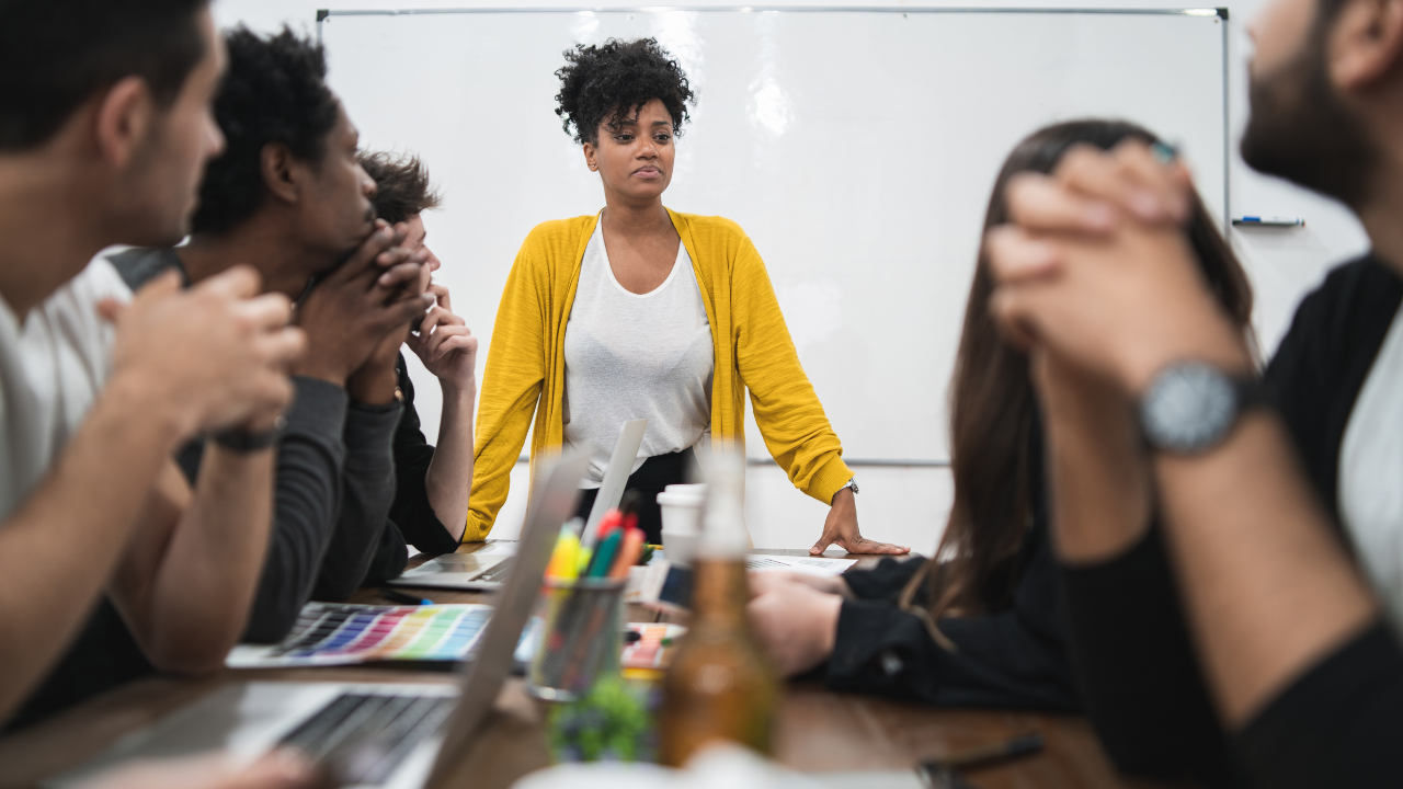 Team members listen as a woman leads a workplace meeting, showing the difference between managing and leading.