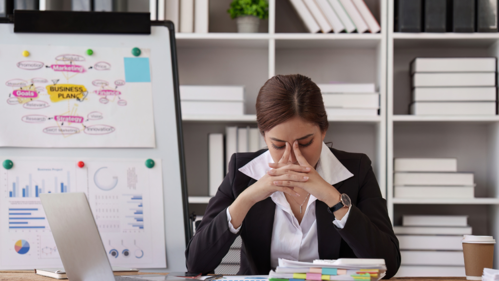 A stressed businesswoman sits at her desk with her head in her hands, surrounded by reports, planning notes, and office materials as her business feels chaotic.