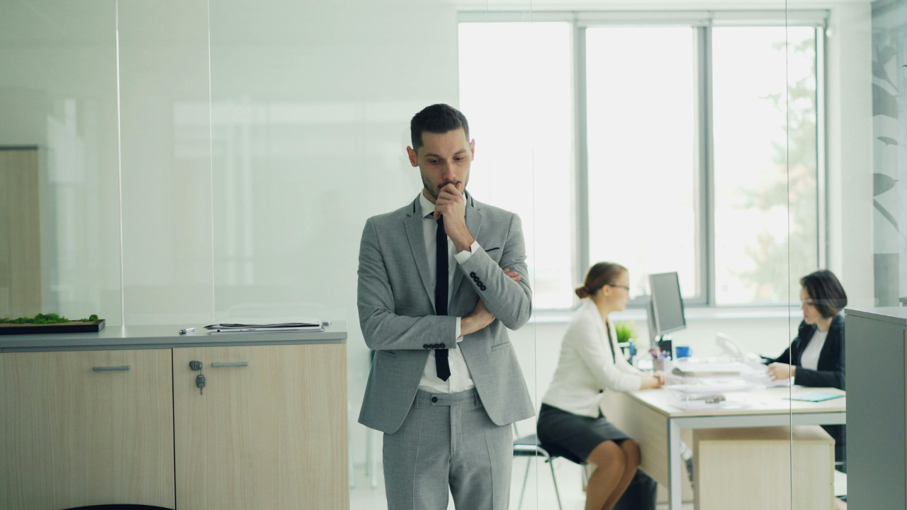 Business leader pausing thoughtfully in a modern office, reflecting how delaying decisions can slow progress while colleagues continue working in the background.