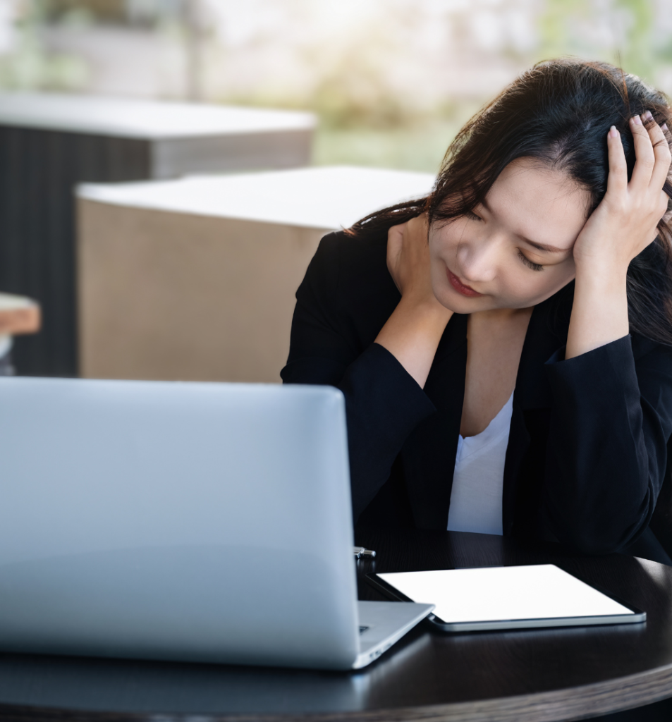 A stressed businesswoman sits at a table with a laptop and tablet, highlighting the challenges of poor operational efficiency in the workplace.