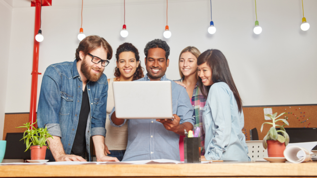 Group of coworkers smiling around a laptop in a bright office, showing team ownership through shared focus and collaboration.
