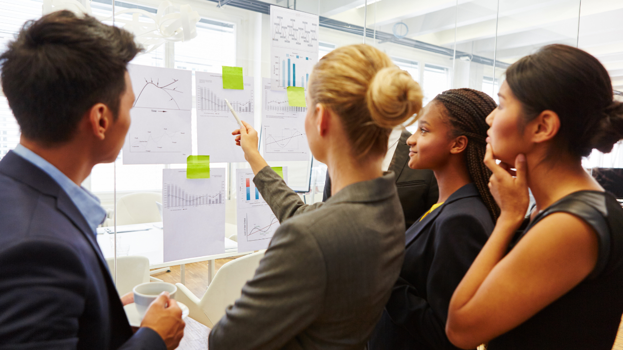 Business forecasting team reviewing charts and performance data on a glass wall during a collaborative office planning meeting.