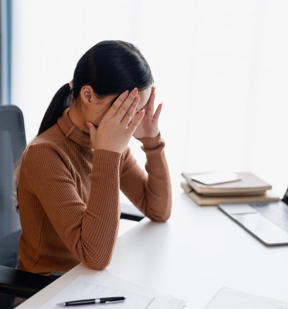 Founder burnout shown as a business owner sitting at her desk with her face in her hands, appearing stressed and exhausted beside a laptop and paperwork.