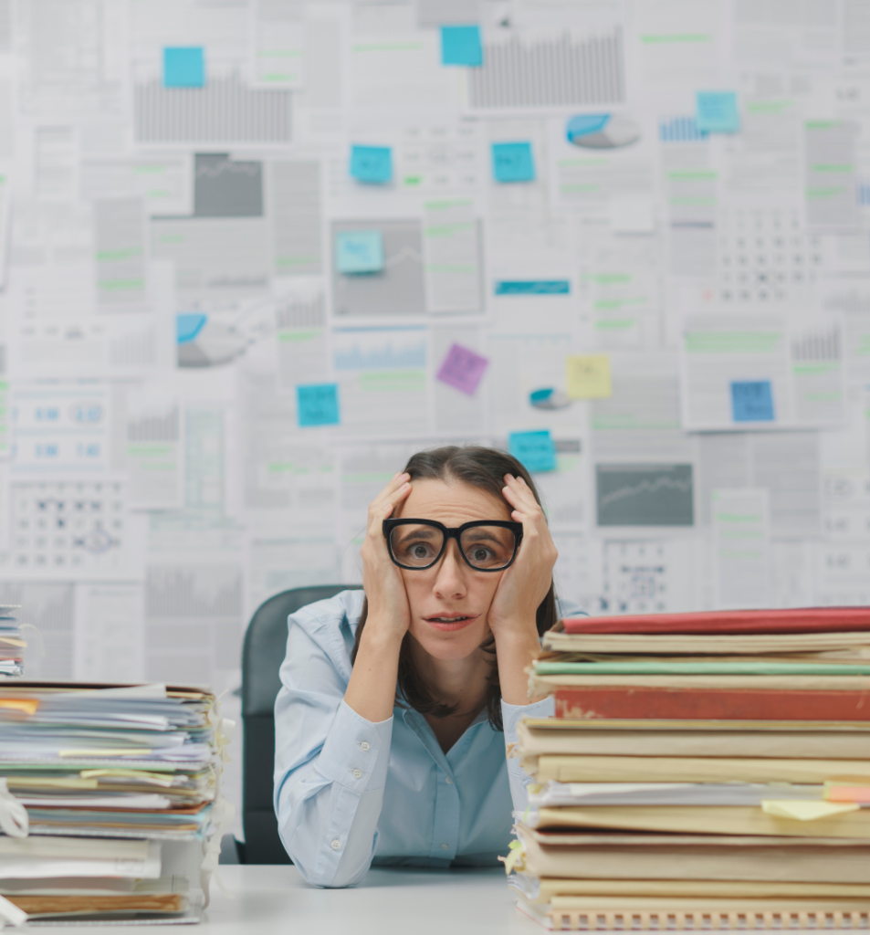 A stressed woman sits at a desk between stacks of paperwork with charts and notes behind her, illustrating leadership overload and the strain of managing too many responsibilities.